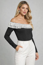 Woman wearing a black off-shoulder top with silver trim and white pants on a gray background
