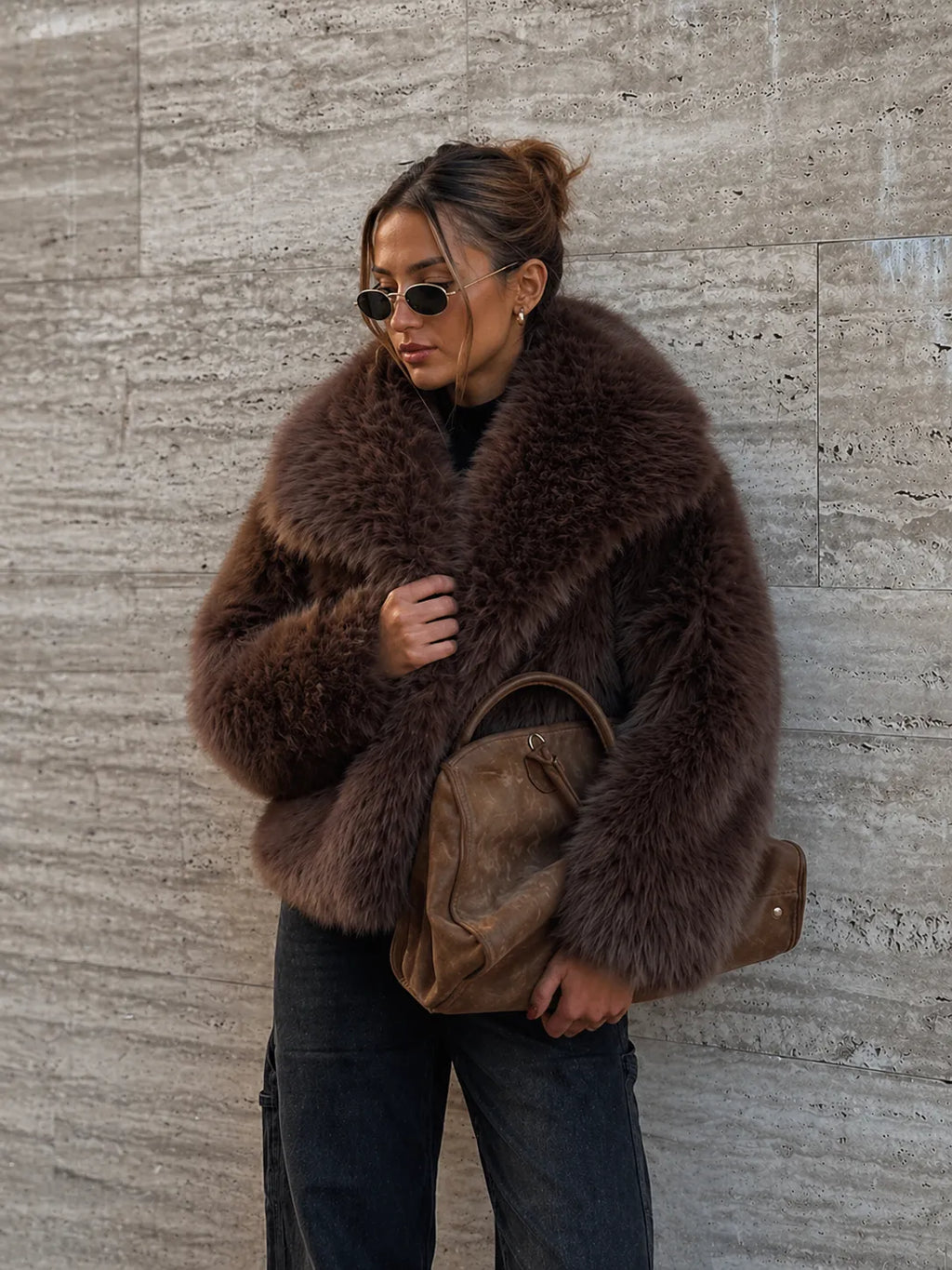 Person wearing a brown fur coat and holding a brown bag against a textured wall.