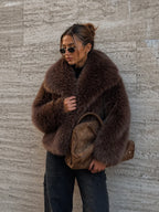 Person wearing a brown fur coat and holding a brown bag against a textured wall.